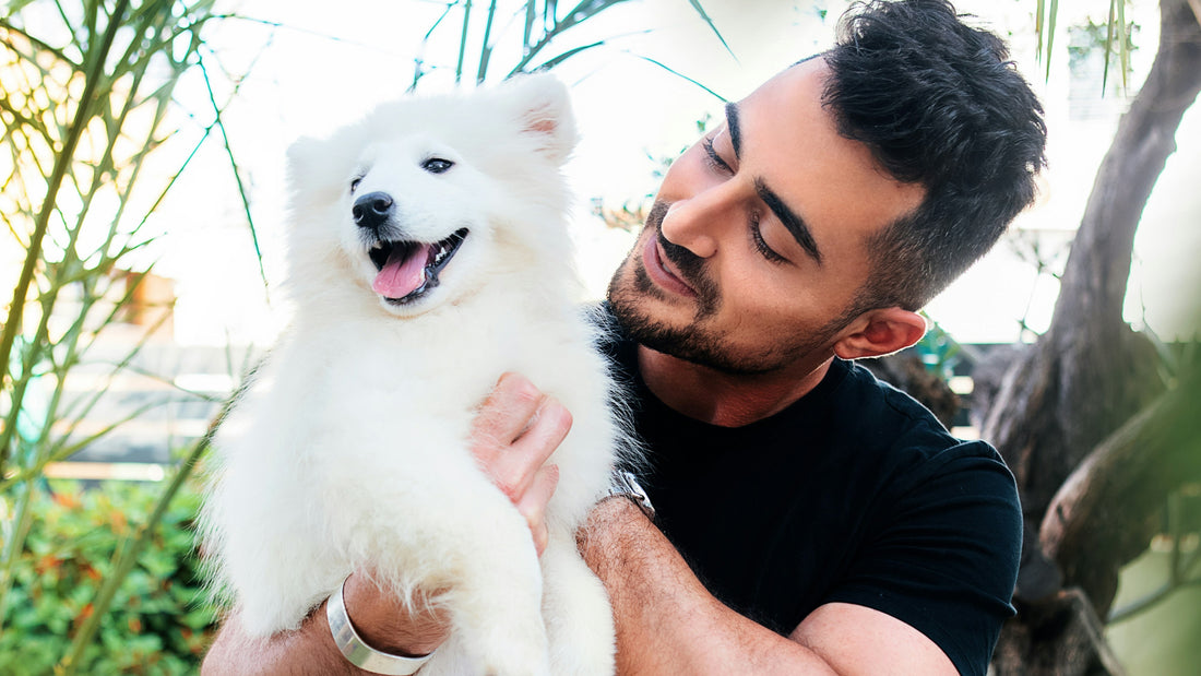 A man in a dark tshirt holds a small white fluffy dog outside in nauture
